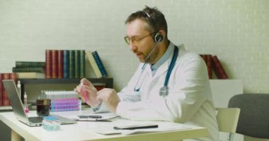 Doktor çevrimiçi video görüşmesi sırasında en yeni aşıdan bahsediyor. Test tüpünde bir dizüstü bilgisayara yenilikçi bir aşı örneği gösteriyor ve keşfinden bahsederken aktif olarak el kol hareketi yapıyor..