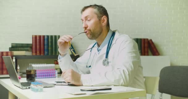 Лікар середнього віку думає через звіт про дослідження, сидячи в клінічному кабінеті . — стокове відео