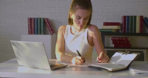 Vista frontal de una joven que trabaja en una oficina: toma notas en papel e introduce datos en una computadora portátil. — Vídeos de Stock
