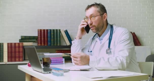 La inyección de Dolyy de un médico de mediana edad está hablando con un paciente por teléfono. Él le habla de los detalles de su diagnóstico y seguro. — Vídeos de Stock