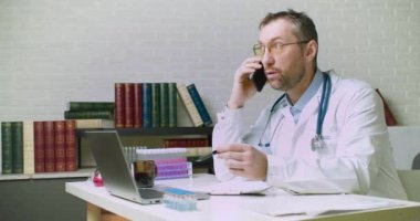 Orta yaşlı bir doktor telefonda bir hastayla konuşuyor. Teşhisinin ve sigortasının detaylarını anlatıyor..