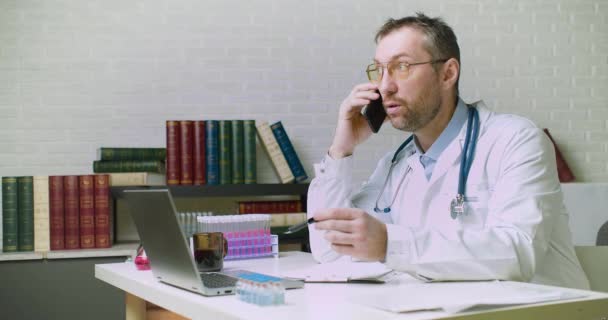 Un médico de mediana edad está hablando con un paciente por teléfono. Él le habla de los detalles de su diagnóstico y seguro. — Vídeos de Stock