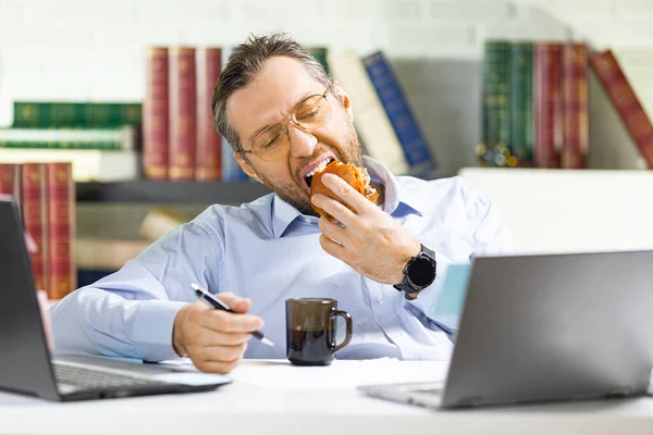 Mann mittleren Alters liegt im Büro - er isst ungesundes Essen. — Stockfoto