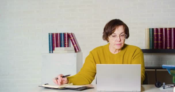 Μια ηλικιωμένη λευκή γυναίκα με γυαλιά που εργάζεται μπροστά από μια οθόνη laptop - ελέγχει την έκθεση και κρατά σημειώσεις στο σημειωματάριό της. — Αρχείο Βίντεο