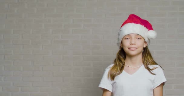Tini lány Santa Hat tartja a karácsonyi ajándékot a kezében egy fehér fal. — Stock videók