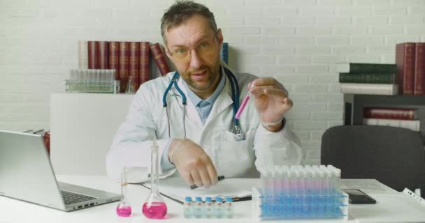 Läkaren pratar om det nyaste vaccinet under ett videosamtal online. Han visar ett vaccin prov i en kamera och aktivt gester när han talar om upptäckter. — Stockvideo