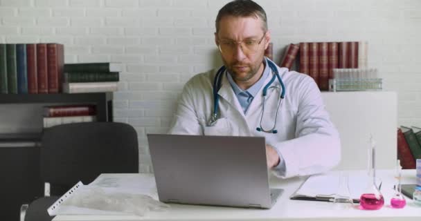 Médico cansado na frente do monitor do laptop ele tira os óculos e esfrega a ponte do nariz. — Vídeo de Stock