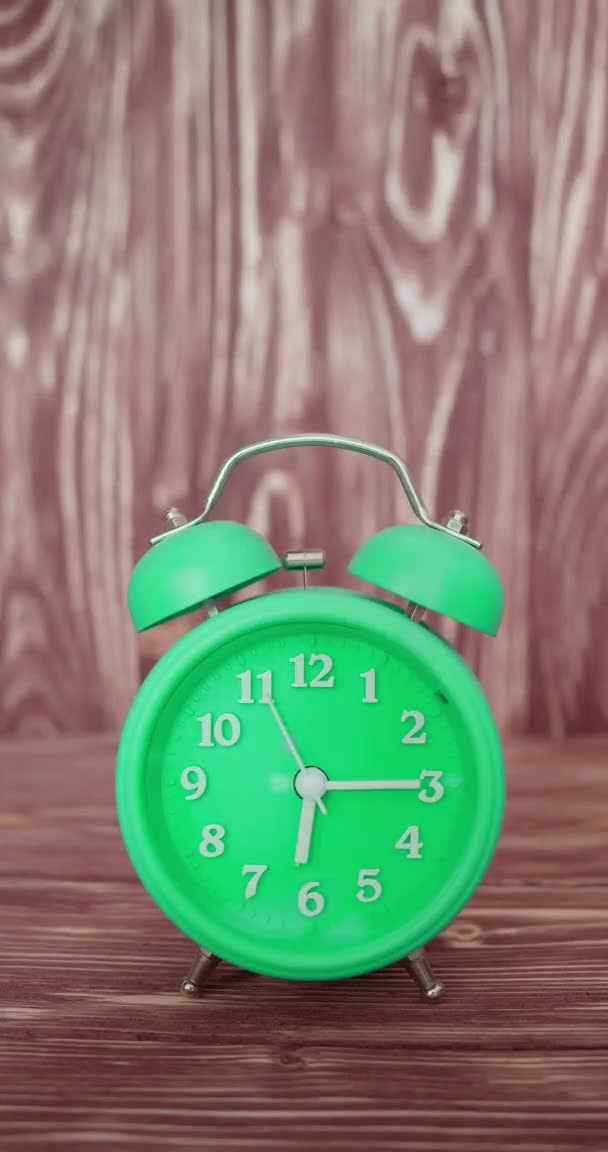 Vertikales Video mit Ton - ein grüner Wecker auf einem hölzernen Hintergrund läutet am frühen Morgen. — Stockvideo