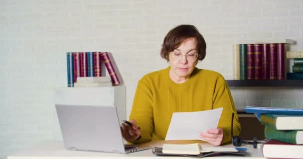 Een oudere blanke vrouw met een bril werkt voor een laptop monitor - ze controleert de belastingformulieren en voert gegevens in in de computer. — Stockvideo