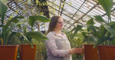 Serada gözlüklü genç bir kadın masaya çiçek koyar, kameraya bakar ve gülümser..