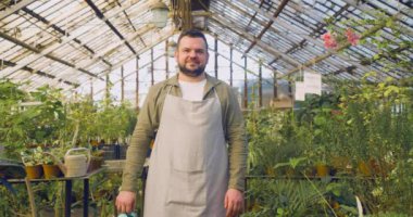 Serada çalışan bir adam. Kameraya bakıyor, gülümsüyor ve elinde bir su kovası tutuyor..