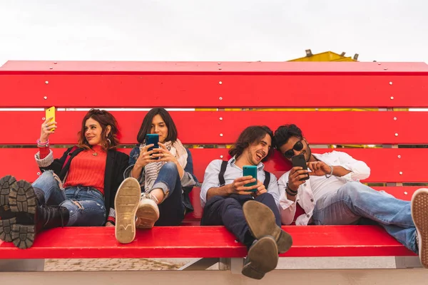 一群多文化的好朋友坐在红色的大长椅上 在户外使用智能手机 — 图库照片
