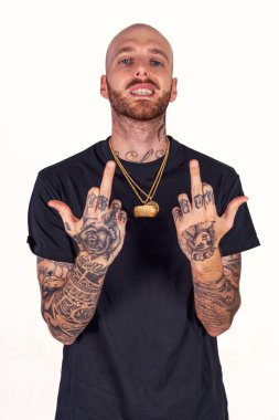 Dövmeli rap şarkıcısı. Stüdyoda poz veriyor. Beyaz arka planda siyah kıyafetler giyiyor.