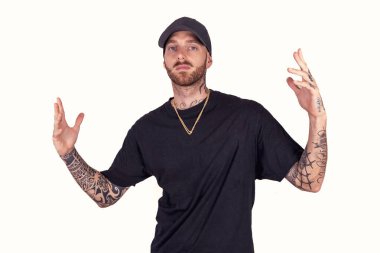 Dövmeli rap şarkıcısı. Stüdyoda poz veriyor. Beyaz arka planda siyah kıyafetler giyiyor.