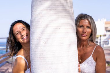 portrait of two beautiful middle aged girlfriends posing by the sea near a white palm tree
