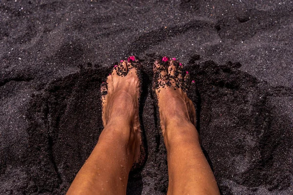 closeup of feet of woman lying down on a black volcanic tropical beach - summer vacation concept
