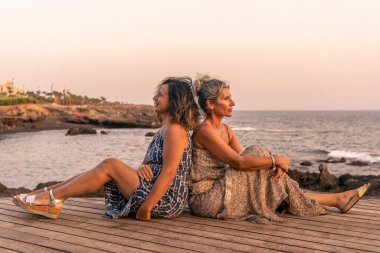 two lovely middle aged girlfriends sitting on a wooden bridge by the sea at sunset - summer vacation concept