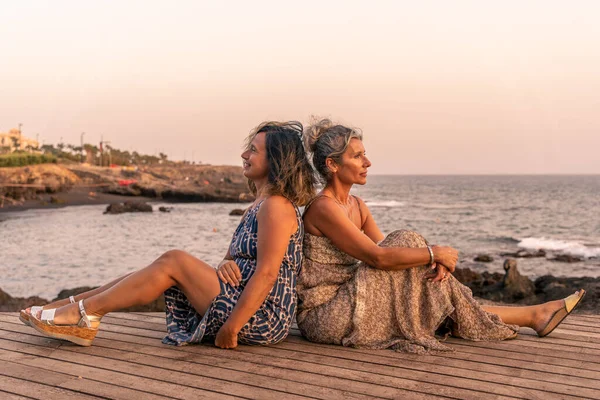 two lovely middle aged girlfriends sitting on a wooden bridge by the sea at sunset - summer vacation concept
