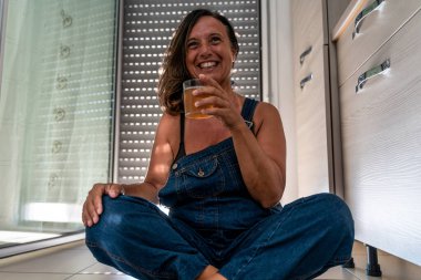 image of a beautiful middle aged woman drinking a cocktail sitting in her apartment - Mature woman dressed a dungarees in his home - Concept about body positivity self esteem and body acceptance