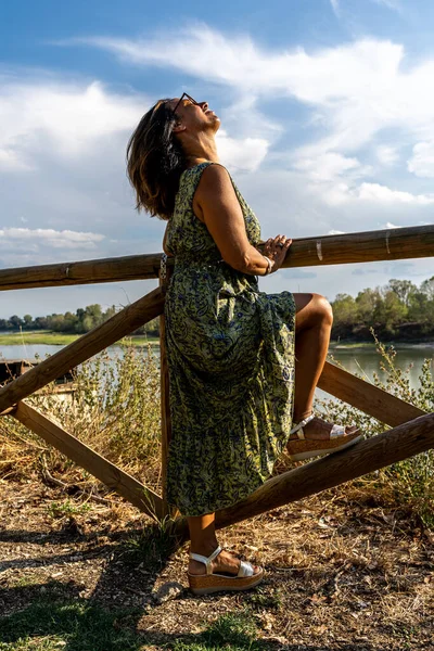 image of beautiful middle aged lady posing leaning against a fence on the banks of the river at sunset