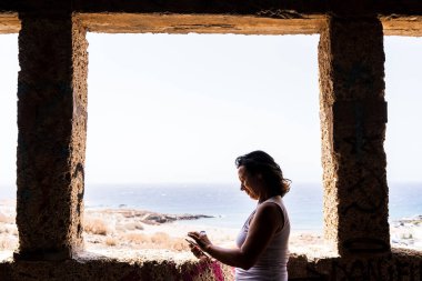 beautiful middle aged woman using phone in front of an open stone window overlooking the sea