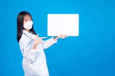 Koruyucu sağlık konsepti. Meslek doktoru ya da hemşire kadın tıbbi ceket giyer ve steteskopla koruyucu yüz maskesi takar. Mavi arka planda duran beyaz tahtayı işaret eder..