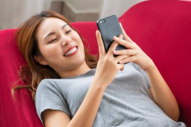 Yaşam tarzı teknoloji konsepti. Oturma odasındaki kırmızı kanepede uzanırken akıllı telefon tutan neşeli genç kadın. Sosyal medyada cep telefonuyla mesaj bağlayan sıradan bir kız..