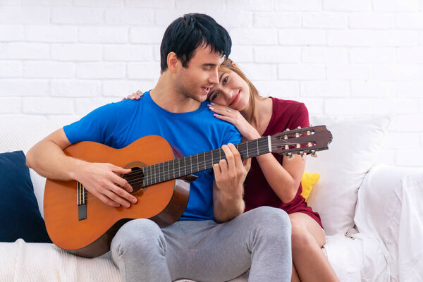 Lifestyle relationship or affectionate concept. Cheerful casual couple playing acoustic guitar leisure while sitting together on white sofa at home. Handsome man singing music for own girlfriend.