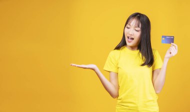 Ödeme ve finansal konsept. Neşeli genç bir kadın, günlük kıyafetler giyip elinde kredi kartı maketi tutarken izole edilmiş sarı arka planda fotokopi aletiyle dikiliyor.. 