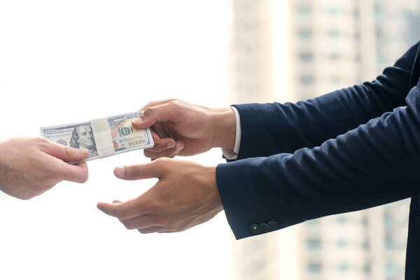 Финансовая концепция оплаты. Рука бизнесмена, держащего в руках долларовые купюры, чтобы оплатить взятку или дать партнеру за размытый городской фон. Селективный фокус 