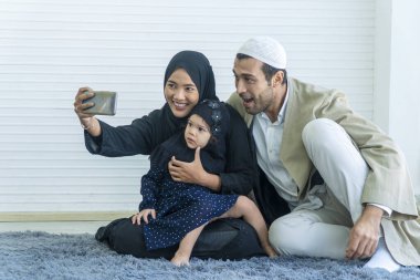 Neşeli Müslüman aile evde birlikte fotoğraf çektirirken cep telefonu kullanıyor. Kızının akıllı telefondan selfie çekip birlikte oturmasının keyfini çıkarmak. Annelik tesettüre girip fotoğraf çektiriyor..