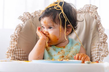 Sevimli, dağınık, küçük bir kız evdeki yüksek güçlü sandalyede oturan el emeği ile spagetti yiyor. Domates soslu küçük bir çocuk, ailesine bakarken yüzünü dağıtıyor. Kendini besleme konsepti
