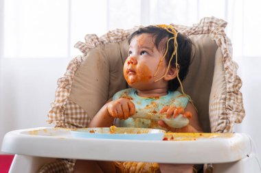 Evdeki güçlü sandalyede oturmuş spagetti kaşığı yiyen sevimli, dağınık, küçük bir kız çocuğu. Domates soslu küçük çocuk ebeveynlerine bakarken yüzünü kirletiyor. Kendini besleme konsepti