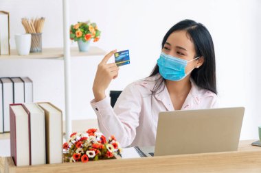 Karantina altındaki Asyalı kadın elinde dizüstü bilgisayarla evde sipariş veriyor. Yüz maskesi takan genç bir kız model kartını gösteriyor. Çevrimiçi e-ticaret ödemesi alışverişi yeni normal konsept