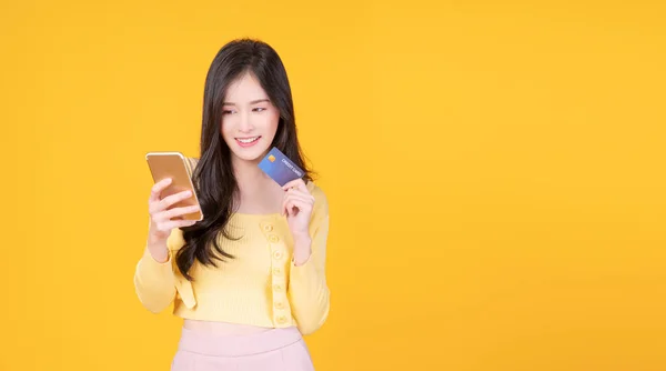 Neşeli gülümseyen Asyalı kadın internet bankacılığı için kredi kartı modeli ve cep telefonu tutuyor izole edilmiş sarı arka planda duruyor. Finansal ödeme çevrimiçi alışveriş ve para transferi kavramı.