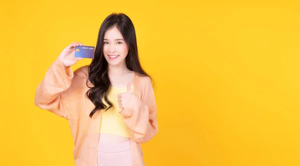 快乐的亚洲年轻女子拿着信用卡高举大拇指进行黄色背景的促销购物 微笑亚洲女孩服务卡特别为客户提供服务 避免与他人隔离 — 图库照片