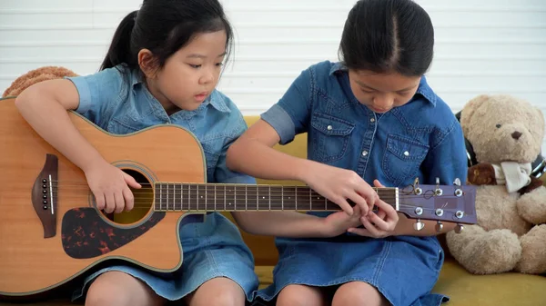 アジアの10代の少女は自宅で黄色のソファに一緒に座ってアコースティックギターを演奏若い教える 幸せな2人の兄弟はリビングでスキルギターを一緒に学ぶ 趣味家の概念 — ストック写真