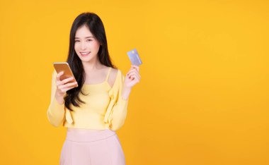 Neşeli gülümseyen Asyalı kadın elinde akıllı telefon ve internet bankacılığı için sahte kredi kartı tutarken izole edilmiş sarı arka planda duruyor. Finansal ödeme çevrimiçi alışveriş ve para transferi kavramı.