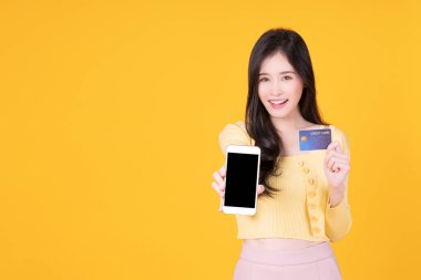 Cheerful smile asian woman showing credit card mockup and mobile phone for internet banking standing over isolated yellow background. Financial payment online shopping and money transfer concept.