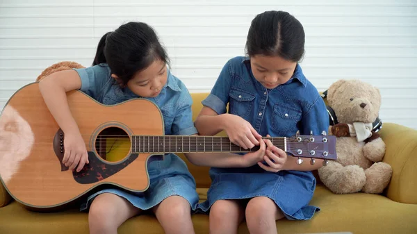 Asian teenage girl teach younger playing acoustic guitar sitting together on yellow sofa at home. Happy two siblings learning play skill guitar togetherness in living room. Hobby family concept.