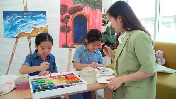 Asian two siblings learning study watercolor paint together at home. Teacher or parent teach student or daughter drawing paint color imagination on paper homework.Activity creativity education concept