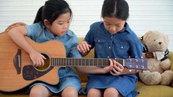 Asian teenage girl teach younger playing acoustic guitar sitting together on yellow sofa at home. Happy two siblings learning play skill guitar togetherness in living room. Hobby family concept.