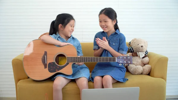 Asian teenage girl teach younger playing acoustic guitar sitting together on yellow sofa at home. Happy two siblings learning play skill guitar togetherness in living room. Hobby family concept.