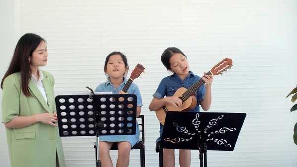Happy Asian two siblings learning play skill guitar togetherness with teacher woman in classroom.Professional teacher training skill hold chords acoustic guitar student.Hobby education musical concept