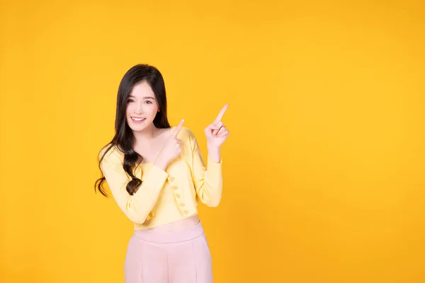 明るいアジアの女性は黄色の背景に隔離された上に立つプロダクトまたは空のコピースペースを指す指を使用する 驚くべきモデルの若い女の子がコンテンツを提示します コンセプトを提示する広告 — ストック写真
