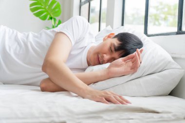Tired asian handsome man sleeping peaceful on white pillow comfortable in morning at apartment with green background. Casual asian young male asleep cozy good dream in bedroom. Lifestyle relax concept