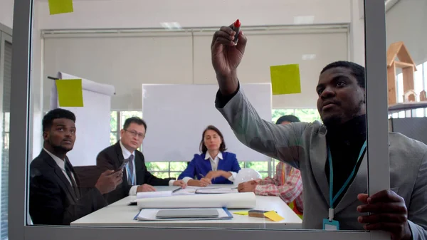 Conference multi-age business people planning project in meeting room at office. African businessman present project write on clear board asian businessman, caucasian businesswoman at workplace.