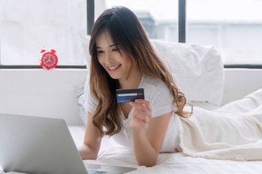 Cheerful asian woman holding credit card or debit mockup electronic payment shopping online with laptop lying on white bed at home. Happy young girl using mockup card pay banking e-commerce.Technology