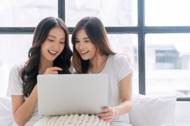Cheerful asian LGBTQ lesbian woman using laptop online sitting together with pillows white bed at apartment. Happy two young girl couple leisure with notebook working online. Technology communication