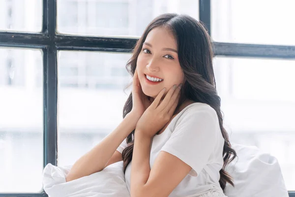 Lovely asian woman casual attire sitting with pillow beside windows relax peaceful in morning at home. Cheerful healthy young girl resting on white pillow with sunlight looking something in bed room.